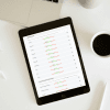 Auf einem weißen Schreibtisch steht neben einem Laptop, schwarzem Kaffee, einem Stift und einer Vase ein Tablet mit einem Leistungs-Dashboard mit Statistiken und Balkendiagrammen sowie Daten zum TAP-Blut-Test Mikronährstoffe.